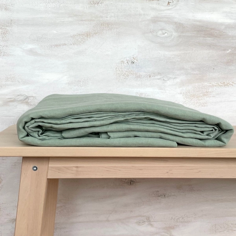 Prześcieradło lniane bez gumki płaskie w kolorze sage green, jasno zielone,  złożone leży na drewnianym stoliku