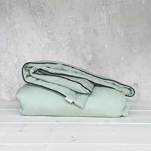 kołdra lniana sage green jasnozielona z czarną lamówką złożona leży na drewnianej jasnej podłodze