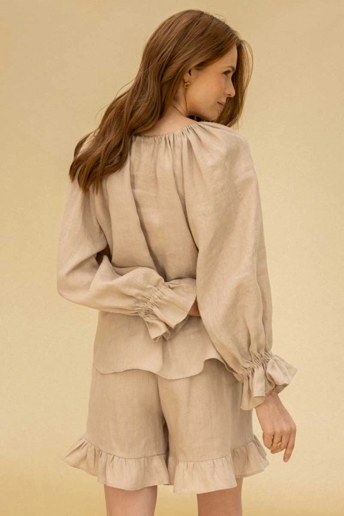 Modelka w lnianej, beżowej bluzce z regulowanym dekoltem i reglanowymi rękawami z falbaną, oraz szortach z falbanami – widok z tyłu. Kolekcja POKOLENIA.