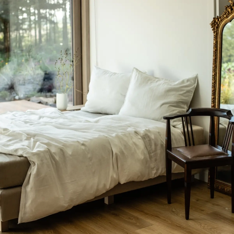 łóżko z lnianą biało kremową pościelą na tle okna z widokiem na las