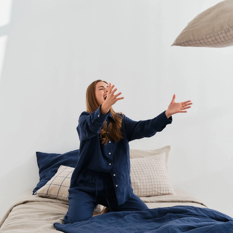 ByMoana23.119340-1.jpg Kobieta w granatowej lnianej piżmie siedzi na łóżku i rzuca poduszkami. Pościel lniana naturalna w granatową kratkę