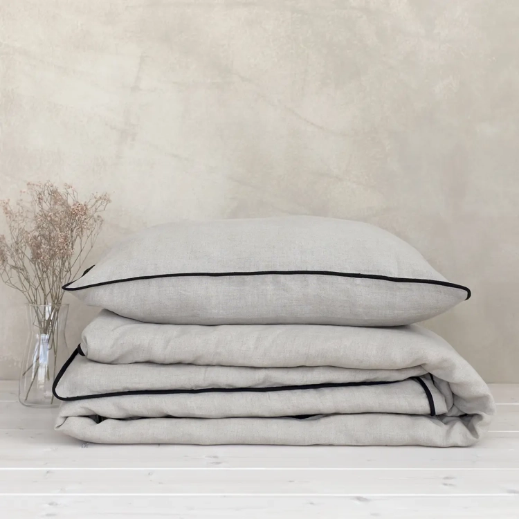 Poduszka leży na złożonej kołdrze. Komplet pościeli lnianej beżowej z czarnym obszyciem